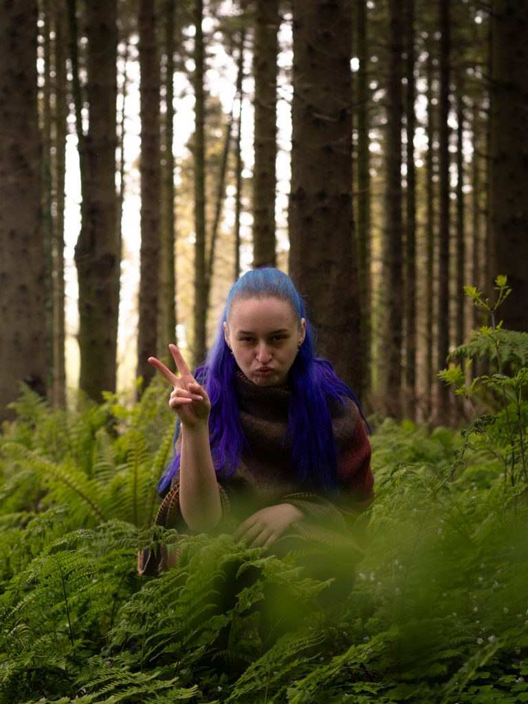 Shannon squatting in the forest giving a peace sign.